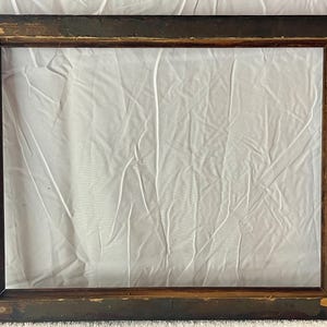 May include: Rectangular wooden picture frame with a dark brown exterior and a lighter brown interior. The frame has visible signs of wear, with some paint chipping off, revealing the wood underneath. The frame is empty, with a white fabric background.