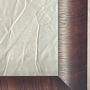 May include: Close-up of a framed artwork. The frame is a dark brown with a wood grain texture. The artwork appears to be a light-colored, textured paper with subtle lines and folds. The corner of the frame is visible.