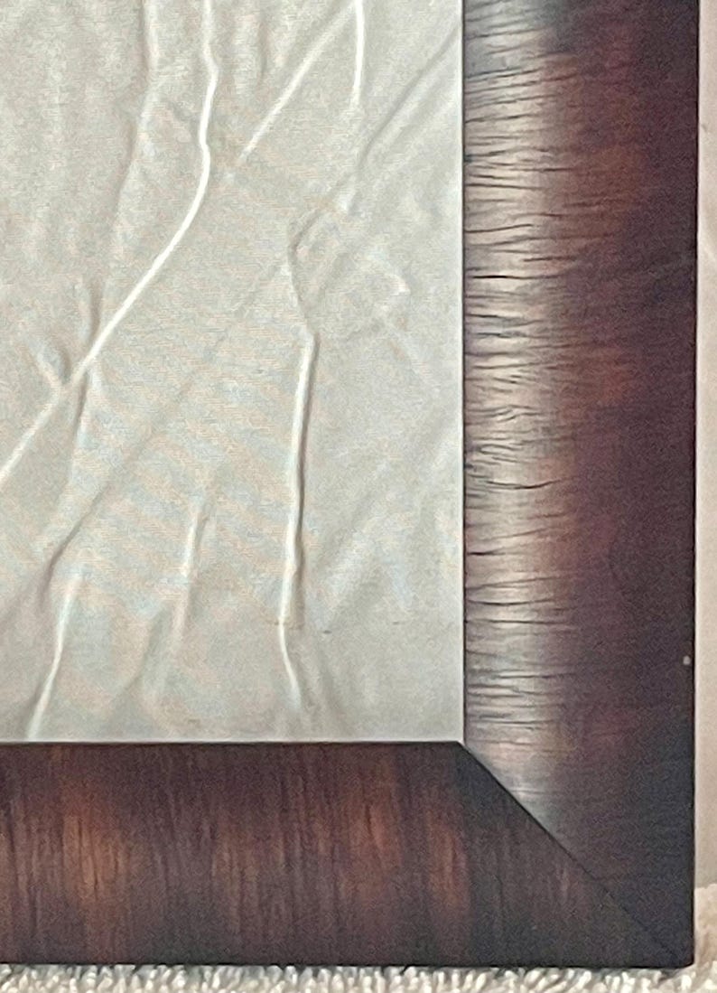 May include: Close-up of a framed artwork. The frame is a dark brown with a wood grain texture. The artwork appears to be a light-colored, textured paper with subtle lines and folds. The corner of the frame is visible.