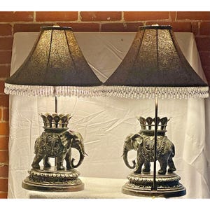 May include: Pair of decorative table lamps. Each lamp features a detailed elephant base with a crown, supporting a black lampshade with a patterned design and crystal fringe. The lamps are approximately 28 inches tall.