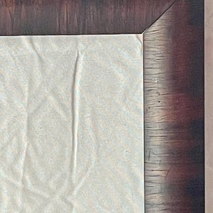 May include: Close-up of a corner of a framed artwork. The frame is a dark brown with a wood grain texture. The artwork is a light-colored, textured fabric.