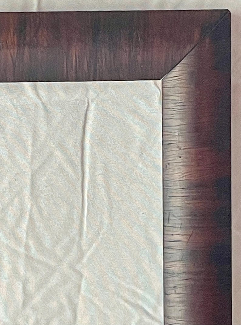 May include: Close-up of a corner of a framed artwork. The frame is a dark brown with a wood grain texture. The artwork is a light-colored, textured fabric.