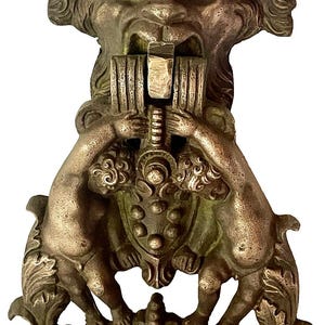 May include: A bronze-toned metal door knocker featuring a central grotesque face with a wide, open mouth. Two small figures are positioned below the face, holding a central shield-like element. The design includes ornate, swirling leaf-like details.