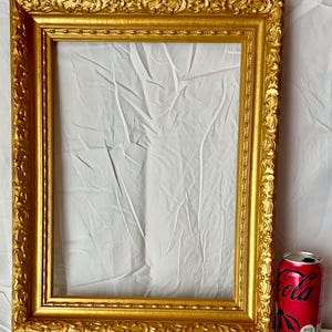 May include: A rectangular, ornate gold picture frame with detailed scrollwork. The frame is empty, with a white fabric backdrop visible. A red can of cola is placed next to the frame for scale.