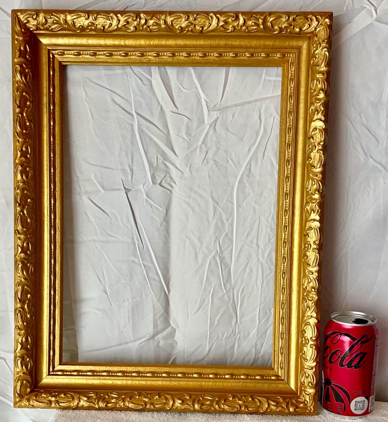 May include: A rectangular, ornate gold picture frame with detailed scrollwork. The frame is empty, with a white fabric backdrop visible. A red can of cola is placed next to the frame for scale.