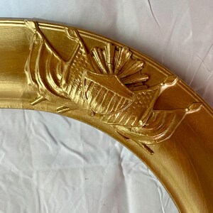 May include: A section of a gold-colored, ornate picture frame. The frame features detailed relief carvings of flags and a sunburst design. The frame has a rounded shape and a glossy finish, suggesting a vintage or antique style.