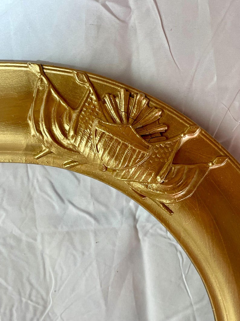 May include: A section of a gold-colored, ornate picture frame. The frame features detailed relief carvings of flags and a sunburst design. The frame has a rounded shape and a glossy finish, suggesting a vintage or antique style.