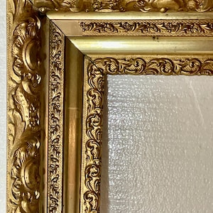 May include: Ornate gold picture frame with detailed carvings. The frame has a rectangular shape with a decorative border and a gold finish. The corners are joined at a 90-degree angle. The frame is empty.