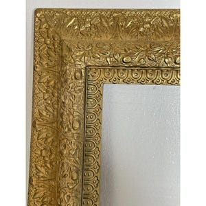 May include: Close-up of a gold-colored picture frame with ornate, embossed floral and leaf detailing. The frame has a rectangular shape with a white interior, and the edges are heavily textured.