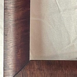 May include: Close-up of a dark brown wooden picture frame with a visible wood grain texture. The frame surrounds a light-colored, possibly fabric, interior. The corner of the frame is visible, showing the angled joinery. The overall aesthetic suggests a classic or vintage style.