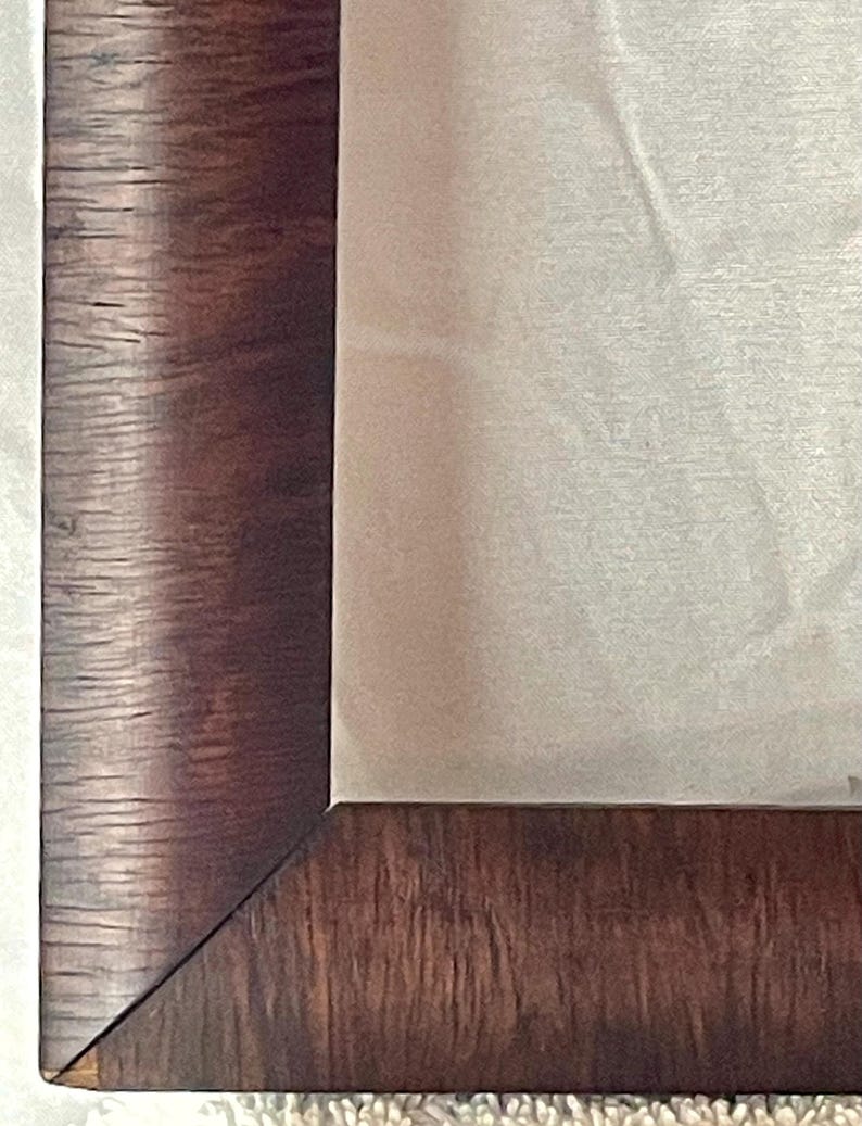 May include: Close-up of a dark brown wooden picture frame with a visible wood grain texture. The frame surrounds a light-colored, possibly fabric, interior. The corner of the frame is visible, showing the angled joinery. The overall aesthetic suggests a classic or vintage style.