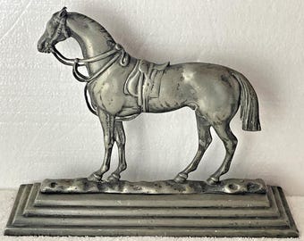 Op de afbeelding: Een zilverkleurig metalen paardenbeeldje op een rechthoekige, getrapte voet. Het paard is in profiel afgebeeld en draagt een zadel en hoofdstel met gedetailleerde teugels. Het oppervlak van het beeldje heeft een matte, verweerde afwerking.