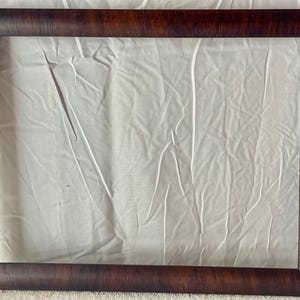 May include: A rectangular, dark brown wooden picture frame with a textured finish. The frame surrounds a white, wrinkled fabric background, creating a contrast in textures and colors. The frame's corners are neatly joined.