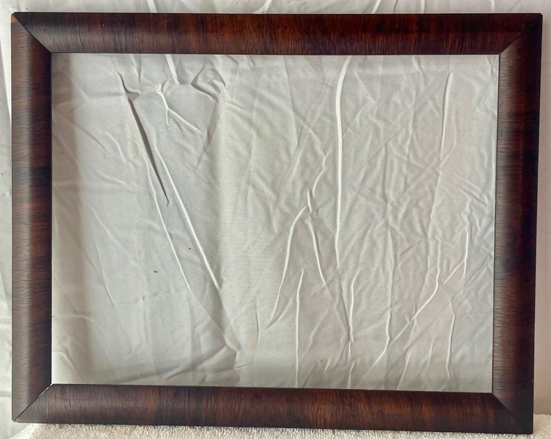 May include: A rectangular, dark brown wooden picture frame with a textured finish. The frame surrounds a white, wrinkled fabric background, creating a contrast in textures and colors. The frame's corners are neatly joined.