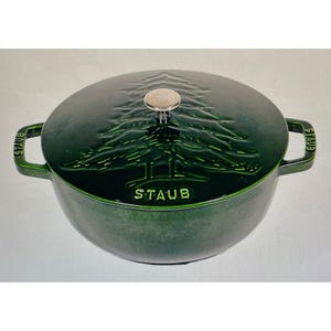 May include: A dark green enameled cast iron Dutch oven with a lid featuring a raised pine tree design. The lid has a silver knob. The word "STAUB" is embossed on the pot and the handles.