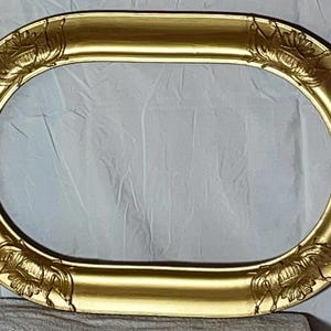 May include: Oval gold picture frame with ornate floral corner details. The frame has a glossy finish and a wide profile. The frame is empty, ready to display a photograph or artwork. The frame is made of a solid material.