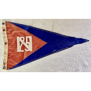 May include: A triangular pennant flag with a blue background and a red diamond shape in the center. The diamond features a white emblem. The flag has a cream-colored border with decorative buttons.
