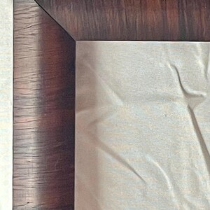 May include: Close-up of a wooden frame corner. The frame has a dark brown, textured finish with visible wood grain. The corner is a precise 90-degree angle. A light-colored, possibly fabric, surface fills the interior space.