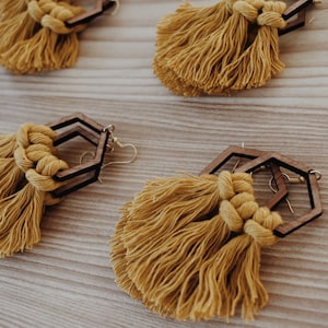 May include: Mustard yellow tassel earrings with geometric wooden frames. The earrings feature a knotted top and gold-colored ear wires. The tassels are made of soft, flowing material. The earrings are displayed on a light-colored wooden surface.
