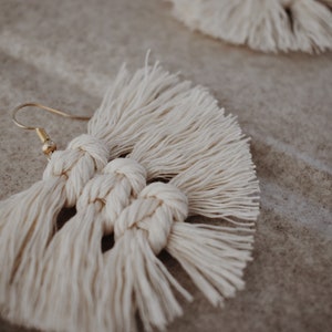 May include: A pair of off-white macrame earrings featuring a fan-shaped design with intricate knotted cord patterns and a gold-toned metal hook. The earrings are crafted from soft cotton-like fiber, showcasing a textured fringe edge and a symmetrical knot structure that hangs from a simple curved wire finding.