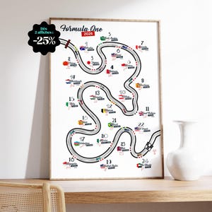 Op de afbeelding: Een ingelijste poster van een Formule 1 2026 racecircuitkaart. Het zwarte circuitontwerp is versierd met vlaggen die verschillende landen vertegenwoordigen. De poster wordt gepresenteerd op een houten oppervlak, naast een witte vaas.