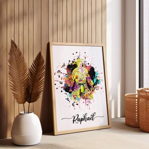 May include: A colorful watercolor print of a person in a yellow martial arts uniform with a black belt. The print is surrounded by colorful paint splatters and the name "Raphael" is written in cursive at the bottom.