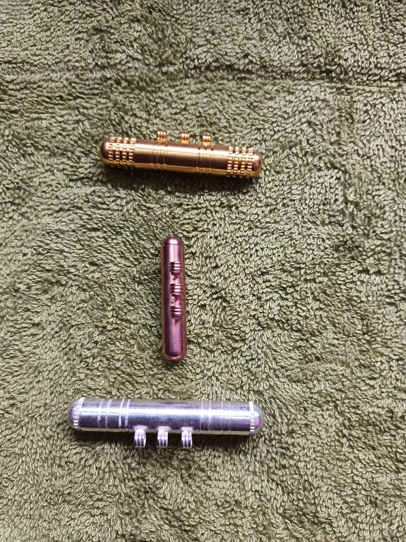 Puede incluir: Tres juguetes de fidget de metal, uno dorado, uno plateado y uno de cobre, sobre una superficie verde texturizada.