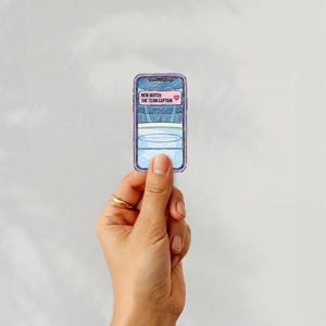 Puede incluir: Una pegatina de un smartphone con un borde morado, sostenida por una mano. La pantalla del tel&eacute;fono muestra el texto "NEW MATCH: THE TEAM CAPTAIN" con un icono de coraz&oacute;n rosa. La pantalla del tel&eacute;fono tiene un dise&ntilde;o azul y blanco.