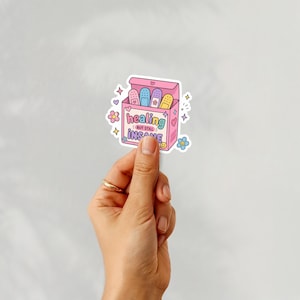 Puede incluir: Una pegatina que representa una caja rosa de tiritas con el texto "healing but still insane". Las tiritas son de varios colores, incluyendo azul, morado y amarillo. La pegatina tiene un estilo de dibujos animados con corazones y flores.