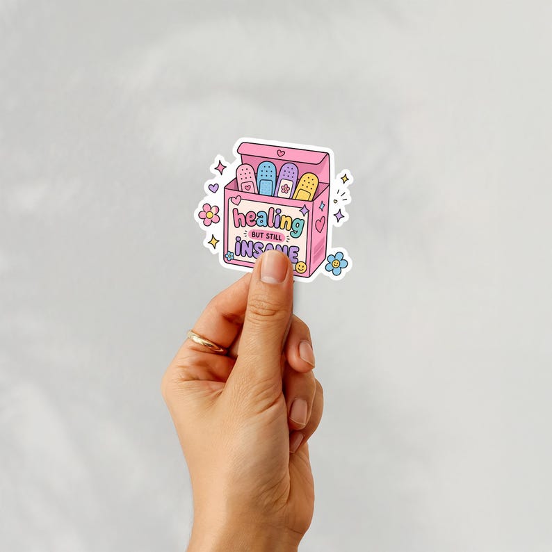 Puede incluir: Una pegatina que representa una caja rosa de tiritas con el texto "healing but still insane". Las tiritas son de varios colores, incluyendo azul, morado y amarillo. La pegatina tiene un estilo de dibujos animados con corazones y flores.