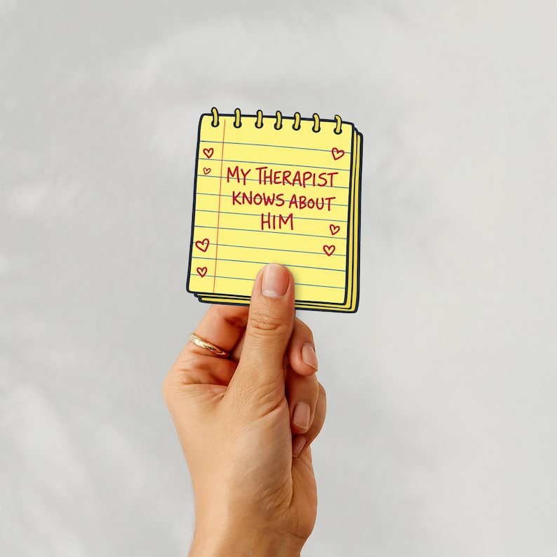 Puede incluir: Un bloc de notas amarillo con el texto "MY THERAPIST KNOWS ABOUT HIM" en negro, rodeado de corazones rojos. El bloc de notas est&aacute; sostenido por una mano con un anillo dorado.