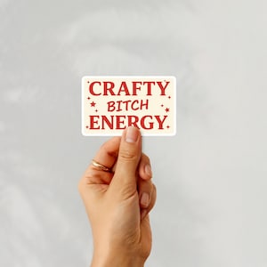 May include: A cream-coloured rectangular sticker with the words "CRAFTY BITCH ENERGY" in red. The sticker has red star accents and is held by a hand wearing a gold ring.