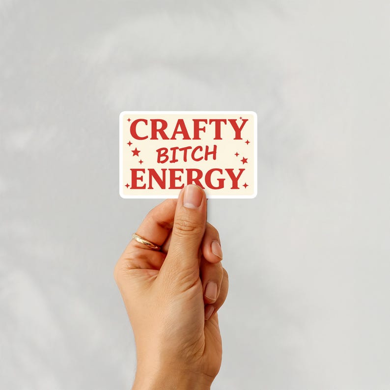 May include: A cream-coloured rectangular sticker with the words "CRAFTY BITCH ENERGY" in red. The sticker has red star accents and is held by a hand wearing a gold ring.
