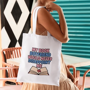 Puede incluir: Bolso tote blanco con el texto "MY BOOK BOYFRIEND WOULD NEVER DISAPPOINT ME" en rosa, azul y negro. El dise&ntilde;o incluye una ilustraci&oacute;n de un libro abierto con dos corazones. El bolso tiene correas blancas.