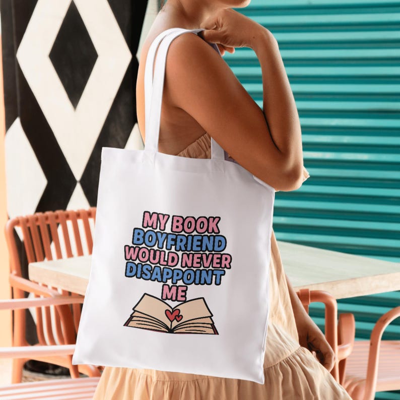 Puede incluir: Bolso tote blanco con el texto "MY BOOK BOYFRIEND WOULD NEVER DISAPPOINT ME" en rosa, azul y negro. El dise&ntilde;o incluye una ilustraci&oacute;n de un libro abierto con dos corazones. El bolso tiene correas blancas.