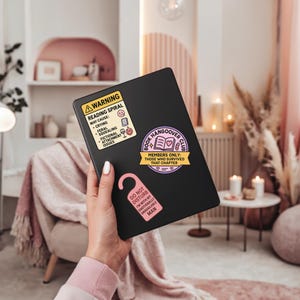 Może przedstawiać: Czarny tablet z trzema naklejkami: ż&oacute;łta etykieta ostrzegawcza o spiralach czytelniczych, r&oacute;żowy wieszak na drzwi i fioletowy emblemat Book Hangover Club. Etykieta ostrzegawcza wymienia potencjalne skutki uboczne.