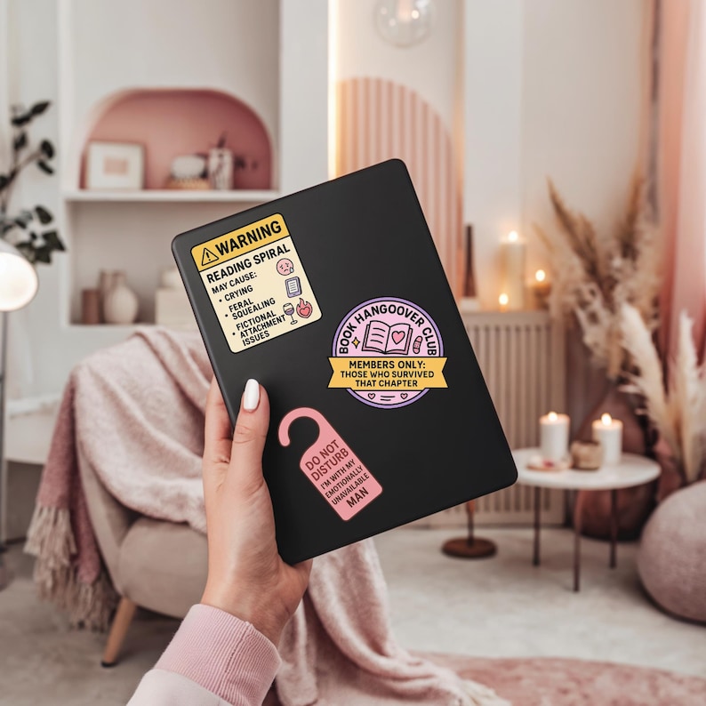 Może przedstawiać: Czarny tablet z trzema naklejkami: ż&oacute;łta etykieta ostrzegawcza o spiralach czytelniczych, r&oacute;żowy wieszak na drzwi i fioletowy emblemat Book Hangover Club. Etykieta ostrzegawcza wymienia potencjalne skutki uboczne.