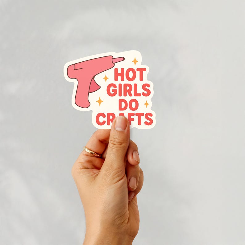May include: A sticker featuring a pink glue gun illustration and the text "HOT GIRLS DO CRAFTS" in red. The sticker has a cream border and is held by a hand with a gold ring.