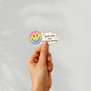 Puede incluir: Una pegatina con una carita sonriente arco&iacute;ris y una burbuja de texto en forma de nube. La burbuja de texto dice "good vibes and emotional damage" en negro. La pegatina se sostiene sobre un fondo blanco liso.