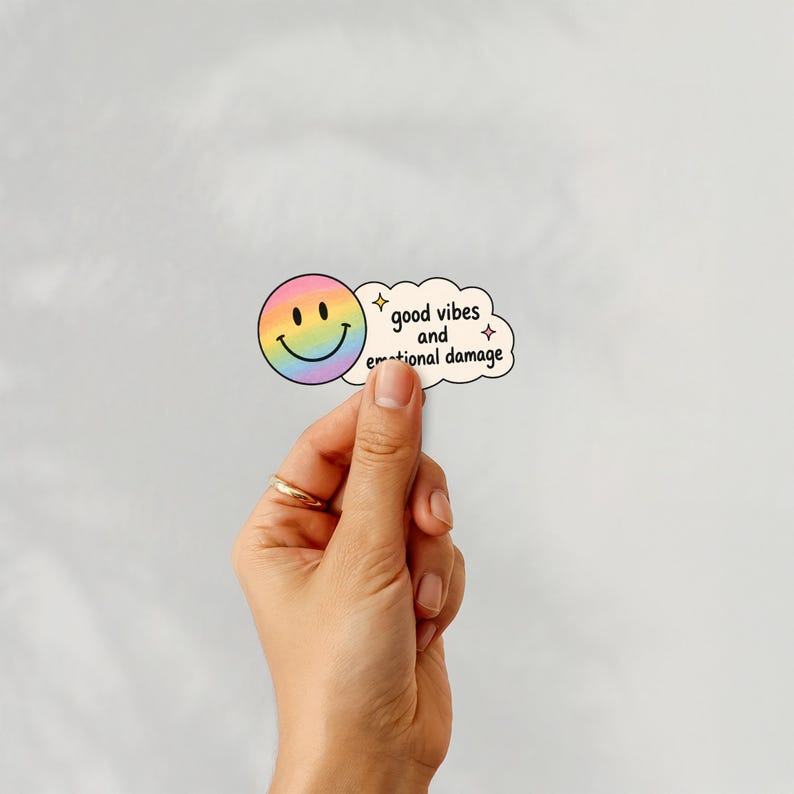 Puede incluir: Una pegatina con una carita sonriente arco&iacute;ris y una burbuja de texto en forma de nube. La burbuja de texto dice "good vibes and emotional damage" en negro. La pegatina se sostiene sobre un fondo blanco liso.