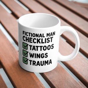 Puede incluir: Taza de cer&aacute;mica blanca con el texto "FICTIONAL MAN CHECKLIST" y las palabras "TATTOOS", "WINGS" y "TRAUMA" debajo. Cada palabra tiene una marca de verificaci&oacute;n verde al lado. La taza est&aacute; sobre una superficie de madera marr&oacute;n.