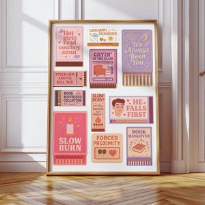May include: A framed print featuring various matchbox-style illustrations with text. The illustrations are in shades of pink, purple, and beige, with phrases like "Hot girls read cowboy smut" and "Slow Burn". The frame is gold, and the background is a white wall and wooden floor.