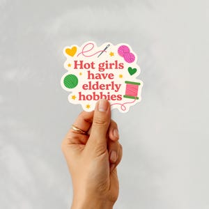 May include: A die-cut sticker with the text "Hot girls have elderly hobbies". The sticker features illustrations of yarn, a needle, and a spool of thread in pink, green, and yellow. The sticker is held by a hand with a gold ring.