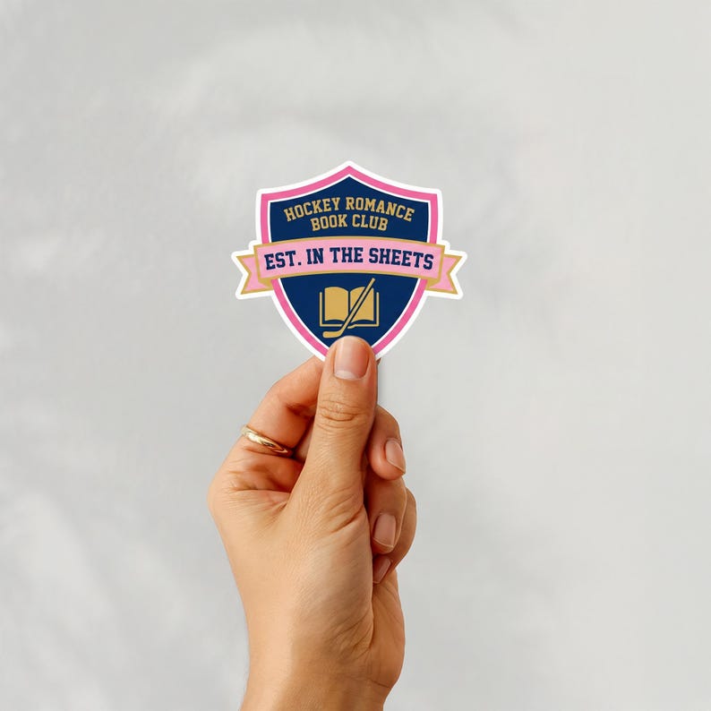 Puede incluir: Una pegatina con un dise&ntilde;o de escudo. El escudo es azul marino con un borde rosa y presenta el texto "HOCKEY ROMANCE BOOK CLUB" y "EST. IN THE SHEETS". Tambi&eacute;n se ven un palo de hockey y un libro. La pegatina est&aacute; sujeta por una mano.