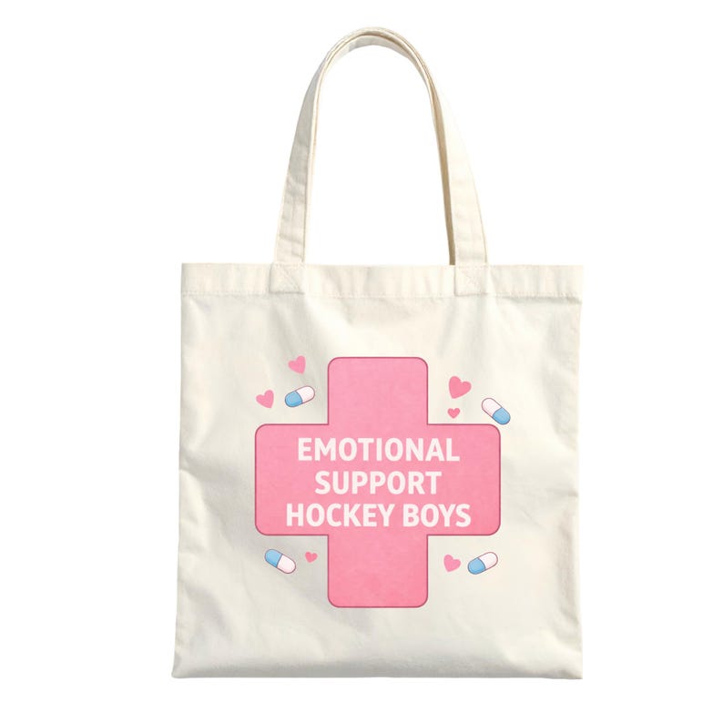 Puede incluir: Bolso tote color crema con un dise&ntilde;o de cruz rosa. La cruz contiene el texto "EMOTIONAL SUPPORT HOCKEY BOYS" en blanco. Peque&ntilde;os corazones rosas y c&aacute;psulas azules y blancas decoran el dise&ntilde;o.