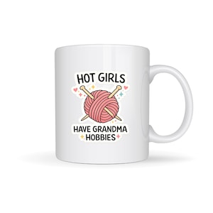 May include: White ceramic mug featuring a pink yarn ball and knitting needles design. The text on the mug reads "HOT GIRLS HAVE GRANDMA HOBBIES". The mug has a curved handle and is suitable for hot or cold drinks.