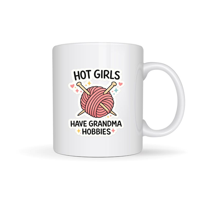 May include: White ceramic mug featuring a pink yarn ball and knitting needles design. The text on the mug reads "HOT GIRLS HAVE GRANDMA HOBBIES". The mug has a curved handle and is suitable for hot or cold drinks.