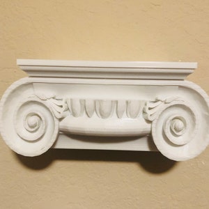 May include: White plaster molding with a decorative scroll design. The molding has a flat top and two scroll designs on the sides. The molding is a decorative element for a home.