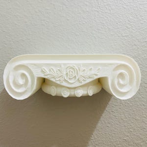 May include: Off-white decorative shelf with a classical column design. The shelf features a floral rose and leaf pattern in the center and scrolled details on each end. The shelf is mounted on a light gray wall.