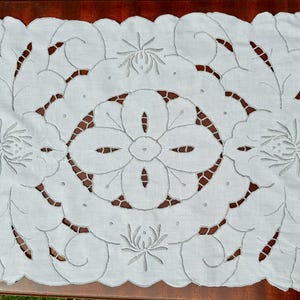 Puede incluir: Camino de mesa rectangular blanco con borde festoneado e intrincados diseños florales y de hojas recortados. El centro presenta una flor grande, rodeada de motivos florales y de hojas más pequeños. El camino de mesa está sobre una superficie marrón oscuro.